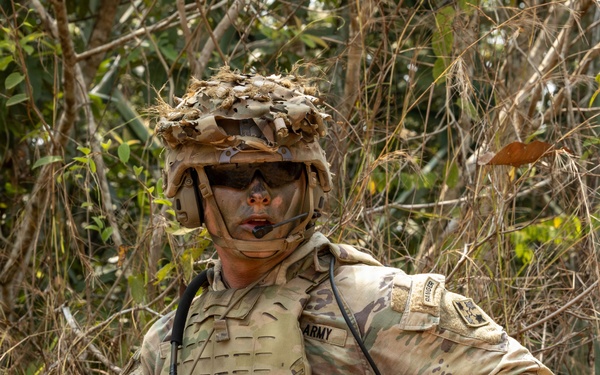 A U.S. Army soldier in full combat gear is positioned in a wooded/brushy outdoor environment. The soldier is wearing MultiCam (OCP) camouflage uniform with a plate carrier vest, tactical helmet with camouflage cover, and face paint. The soldier is holding an M4-style carbine rifle with an optic and appears to be in a field training or operational setting.