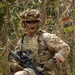 A U.S. Army soldier in full combat gear is positioned in a wooded/brushy outdoor environment. The soldier is wearing MultiCam (OCP) camouflage uniform with a plate carrier vest, tactical helmet with camouflage cover, and face paint. The soldier is holding an M4-style carbine rifle with an optic and appears to be in a field training or operational setting.