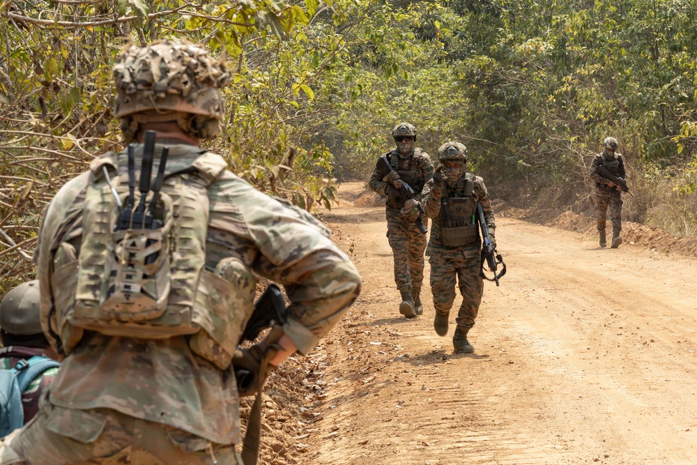 The image shows a group of military personnel conducting a patrol along a dirt road through a wooded or jungle-like terrain. Multiple soldiers in different camouflage patterns are visible, suggesting a multinational or joint training exercise. The foreground soldier is equipped with a tactical backpack and rifle, facing toward the approaching patrol members.