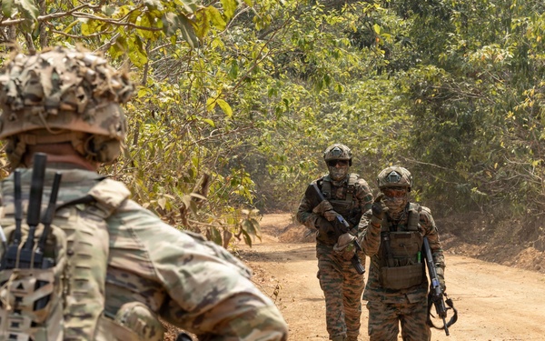 The image shows a group of military personnel conducting a patrol along a dirt road through a wooded or jungle-like terrain. Multiple soldiers in different camouflage patterns are visible, suggesting a multinational or joint training exercise. The foreground soldier is equipped with a tactical backpack and rifle, facing toward the approaching patrol members.