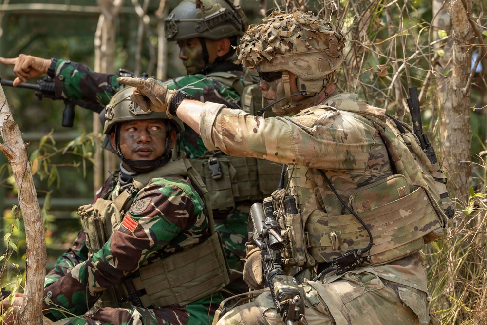 The image shows three soldiers in a wooded outdoor environment, appearing to be engaged in a military training exercise or tactical operation. Two soldiers are wearing Indonesian Army (TNI-AD) camouflage uniforms with distinctive brown-green pattern, while a third soldier wears MultiCam/OCP pattern gear consistent with US Army special operations. The soldier in MultiCam is pointing in a direction, suggesting coordination or target designation between allied forces.