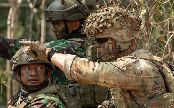 The image shows three soldiers in a wooded outdoor environment, appearing to be engaged in a military training exercise or tactical operation. Two soldiers are wearing Indonesian Army (TNI-AD) camouflage uniforms with distinctive brown-green pattern, while a third soldier wears MultiCam/OCP pattern gear consistent with US Army special operations. The soldier in MultiCam is pointing in a direction, suggesting coordination or target designation between allied forces.