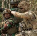 The image shows three soldiers in a wooded outdoor environment, appearing to be engaged in a military training exercise or tactical operation. Two soldiers are wearing Indonesian Army (TNI-AD) camouflage uniforms with distinctive brown-green pattern, while a third soldier wears MultiCam/OCP pattern gear consistent with US Army special operations. The soldier in MultiCam is pointing in a direction, suggesting coordination or target designation between allied forces.