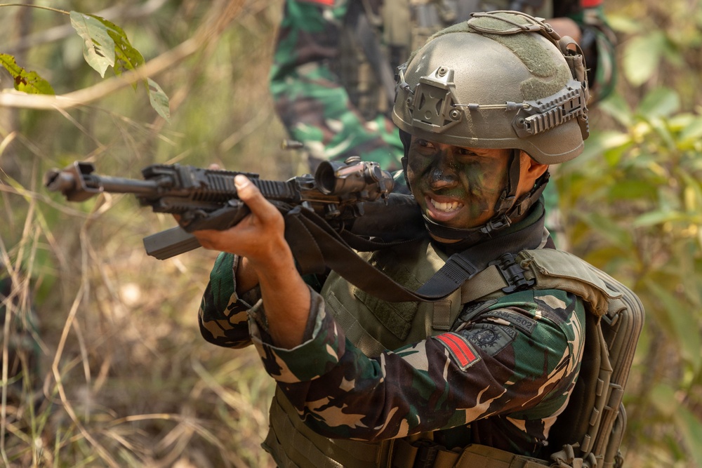 A soldier in full combat gear is shown in a tactical stance aiming a rifle in a jungle or forested environment. The soldier is wearing camouflage face paint, a tactical helmet with rail accessories, body armor, and camouflage uniform with visible unit patches. A second soldier in camouflage uniform is partially visible in the background.