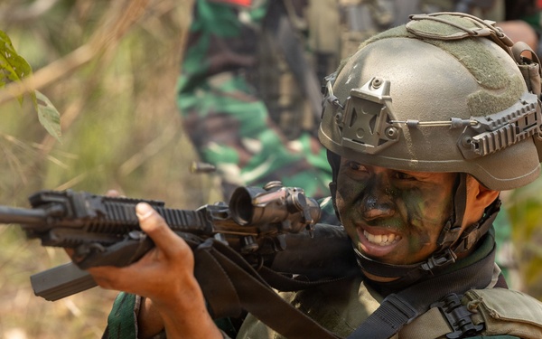 A soldier in full combat gear is shown in a tactical stance aiming a rifle in a jungle or forested environment. The soldier is wearing camouflage face paint, a tactical helmet with rail accessories, body armor, and camouflage uniform with visible unit patches. A second soldier in camouflage uniform is partially visible in the background.