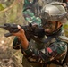 A soldier in full combat gear is shown in a tactical stance aiming a rifle in a jungle or forested environment. The soldier is wearing camouflage face paint, a tactical helmet with rail accessories, body armor, and camouflage uniform with visible unit patches. A second soldier in camouflage uniform is partially visible in the background.