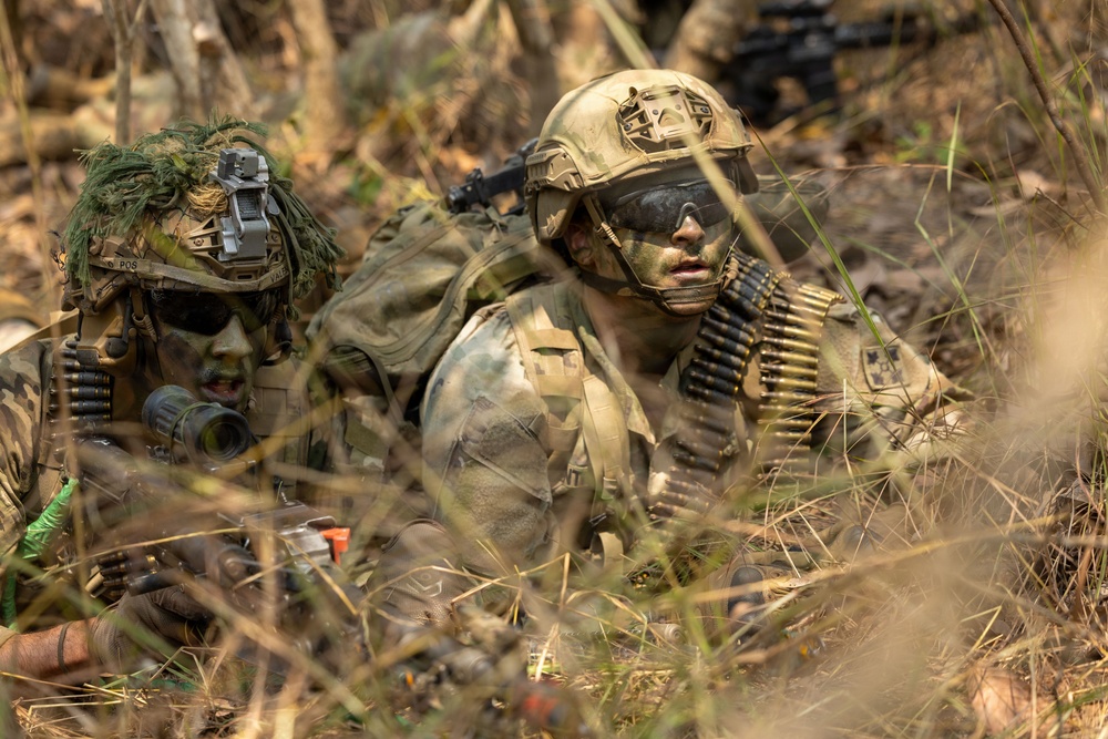 Two heavily camouflaged soldiers are lying prone in a grassy, wooded area in what appears to be a military field exercise or training operation. Both wear full camouflage face paint and tactical gear, with one soldier carrying a belt of linked ammunition across his chest. A third figure is partially visible in the background.