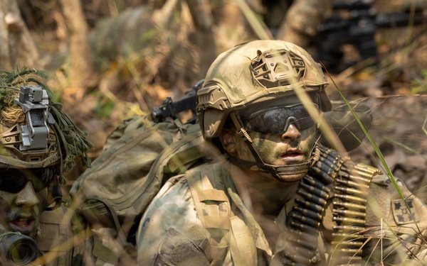 Two heavily camouflaged soldiers are lying prone in a grassy, wooded area in what appears to be a military field exercise or training operation. Both wear full camouflage face paint and tactical gear, with one soldier carrying a belt of linked ammunition across his chest. A third figure is partially visible in the background.