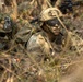 Two heavily camouflaged soldiers are lying prone in a grassy, wooded area in what appears to be a military field exercise or training operation. Both wear full camouflage face paint and tactical gear, with one soldier carrying a belt of linked ammunition across his chest. A third figure is partially visible in the background.