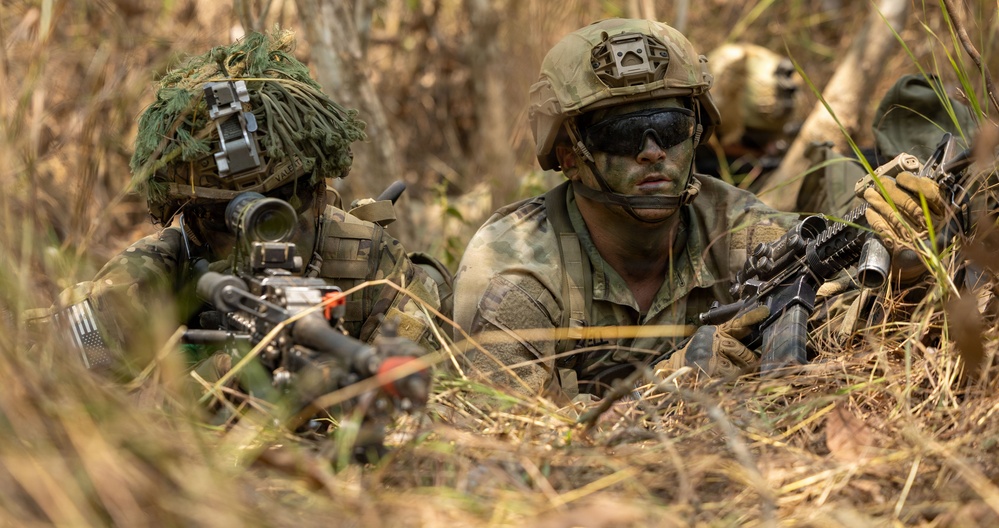 Three military personnel in full combat gear are positioned low in dry grassland/brush terrain, appearing to be in a tactical prone or crouching position. The soldiers are equipped with assault rifles, optical sights, helmets with mounted devices, and camouflage face paint. A US flag patch is visible on one soldier's sleeve, suggesting US military affiliation during what appears to be a field training exercise or operational deployment.