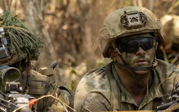 Three military personnel in full combat gear are positioned low in dry grassland/brush terrain, appearing to be in a tactical prone or crouching position. The soldiers are equipped with assault rifles, optical sights, helmets with mounted devices, and camouflage face paint. A US flag patch is visible on one soldier's sleeve, suggesting US military affiliation during what appears to be a field training exercise or operational deployment.