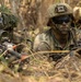 Three military personnel in full combat gear are positioned low in dry grassland/brush terrain, appearing to be in a tactical prone or crouching position. The soldiers are equipped with assault rifles, optical sights, helmets with mounted devices, and camouflage face paint. A US flag patch is visible on one soldier's sleeve, suggesting US military affiliation during what appears to be a field training exercise or operational deployment.