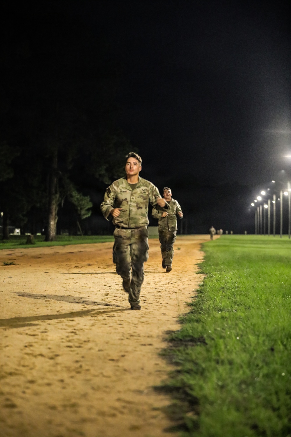 Two military personnel in camouflage uniforms are running along a sandy dirt path at night, illuminated by a row of overhead lights. The lead soldier is clearly visible in the foreground, with a second soldier running behind. The setting appears to be a military installation training area or parade field.