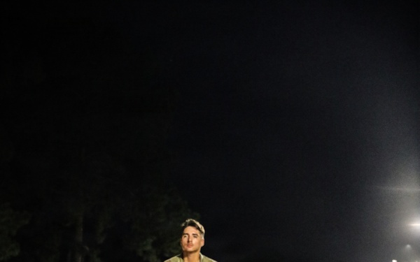 Two military personnel in camouflage uniforms are running along a sandy dirt path at night, illuminated by a row of overhead lights. The lead soldier is clearly visible in the foreground, with a second soldier running behind. The setting appears to be a military installation training area or parade field.