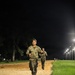 Two military personnel in camouflage uniforms are running along a sandy dirt path at night, illuminated by a row of overhead lights. The lead soldier is clearly visible in the foreground, with a second soldier running behind. The setting appears to be a military installation training area or parade field.