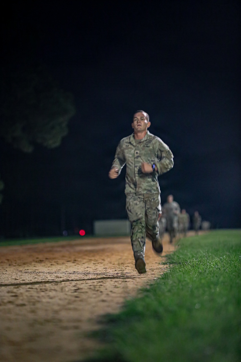 A U.S. Army soldier in OCP (Operational Camouflage Pattern) uniform is running at the front of a formation along a dirt path at night. The soldier appears to be leading a physical training run, with several other uniformed personnel visible in the background. The scene is illuminated by artificial lighting, suggesting a military base or training facility setting.