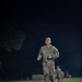 A U.S. Army soldier in OCP (Operational Camouflage Pattern) uniform is running at the front of a formation along a dirt path at night. The soldier appears to be leading a physical training run, with several other uniformed personnel visible in the background. The scene is illuminated by artificial lighting, suggesting a military base or training facility setting.