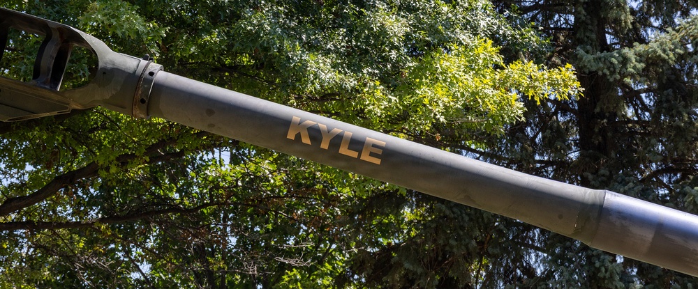 The image shows a large metal barrel or tube with the name 'KYLE' stenciled in gold/yellow lettering on its surface. The object appears to be a cannon or artillery barrel, likely from a military vehicle or static display. It is photographed at an upward angle against a backdrop of green deciduous and coniferous trees.