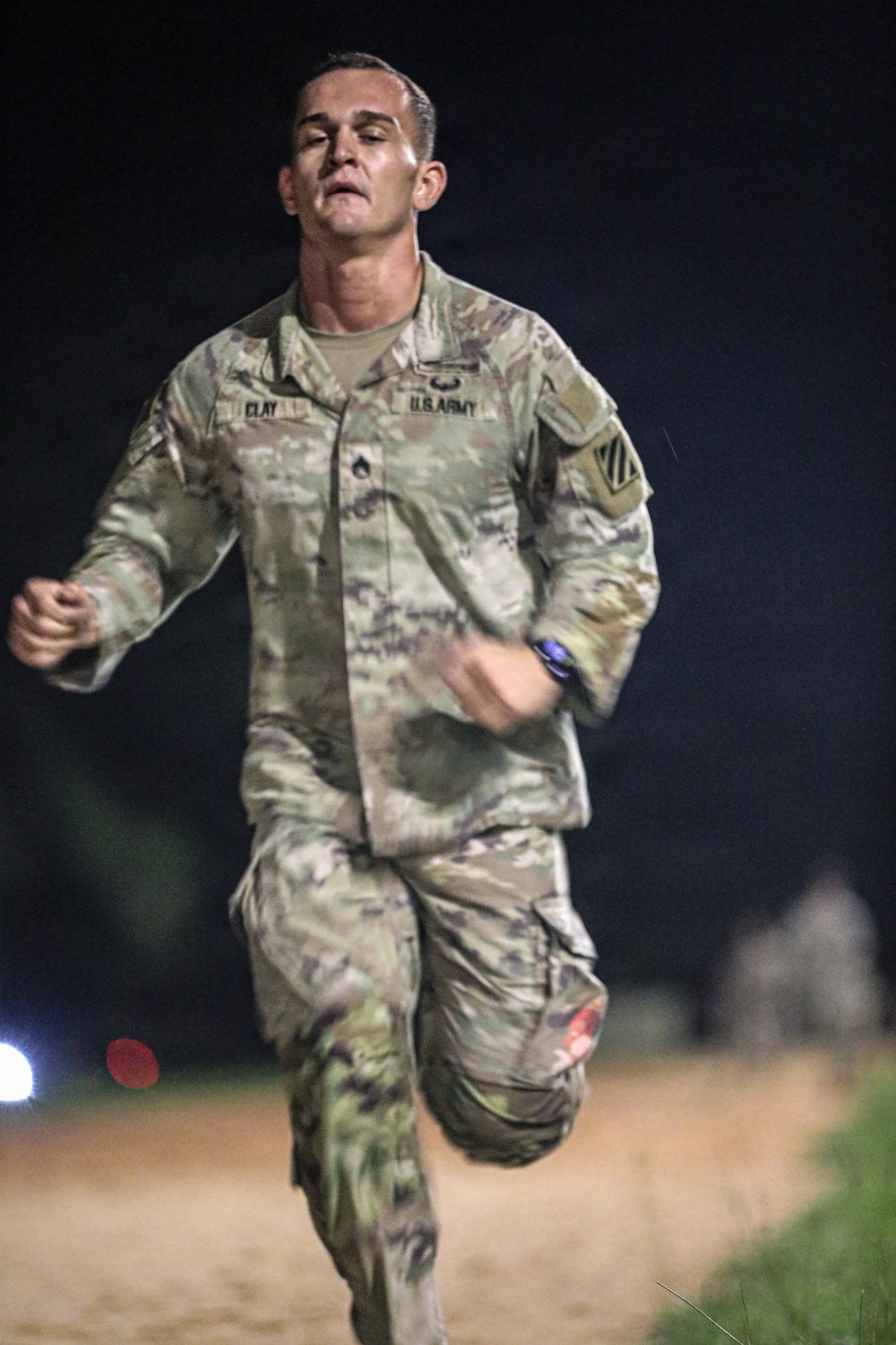 A U.S. Army soldier wearing OCP (Operational Camouflage Pattern) uniform is running at night on what appears to be a dirt path or track. The soldier's name tape reads 'CLAY' and the branch tape reads 'U.S. ARMY,' with a unit patch visible on the left shoulder. The image captures an apparent physical fitness test or training run, with the soldier showing visible exertion.