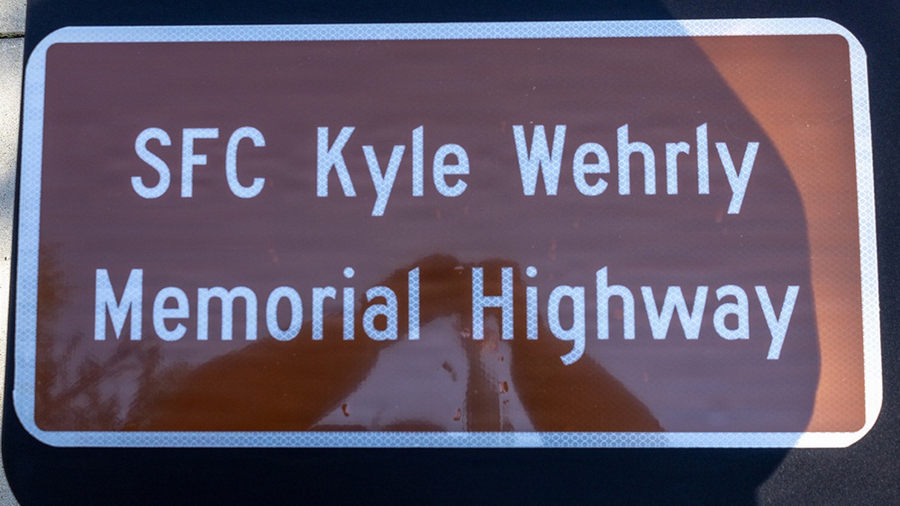 The image shows a brown memorial highway sign with white text and a white reflective border. The sign designates a road as the 'SFC Kyle Wehrly Memorial Highway,' honoring a Sergeant First Class. A shadow or reflection is visible on the sign's surface, likely cast by a nearby object or person.