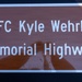 The image shows a brown memorial highway sign with white text and a white reflective border. The sign designates a road as the 'SFC Kyle Wehrly Memorial Highway,' honoring a Sergeant First Class. A shadow or reflection is visible on the sign's surface, likely cast by a nearby object or person.