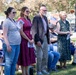 An outdoor public ceremony or memorial event with four people standing in the foreground, appearing to be participants or honorees. An American flag is visible in the background among a seated crowd gathered on a grass lawn. The event appears formal in nature, with some attendees holding printed programs.