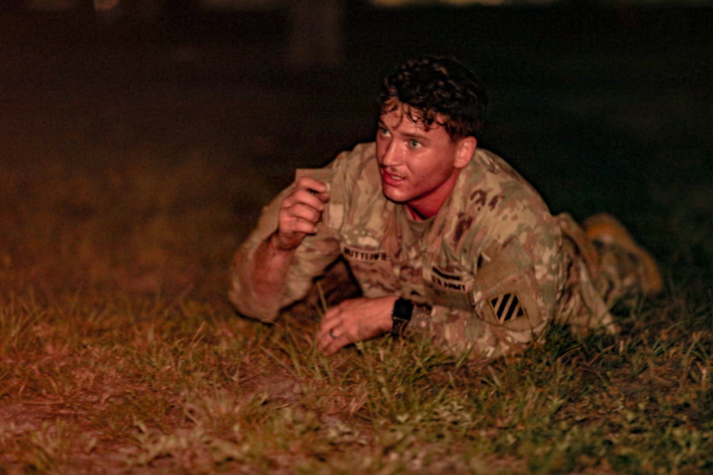 A U.S. Army soldier in OCP (Operational Camouflage Pattern) uniform is low-crawling on grass during what appears to be a nighttime training exercise. The soldier appears physically exhausted and wet, suggesting an intense physical training or obstacle course event. The unit patch on the left shoulder indicates affiliation with the 3rd Infantry Division.