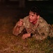 A U.S. Army soldier in OCP (Operational Camouflage Pattern) uniform is low-crawling on grass during what appears to be a nighttime training exercise. The soldier appears physically exhausted and wet, suggesting an intense physical training or obstacle course event. The unit patch on the left shoulder indicates affiliation with the 3rd Infantry Division.