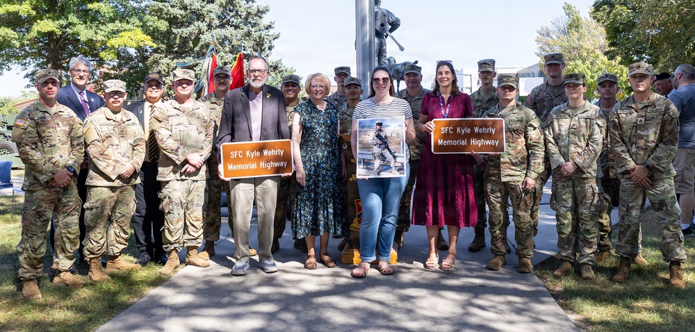 A group of military personnel and civilians gather for a memorial highway dedication ceremony honoring SFC Kyle Wehrly. Two participants hold brown highway signs reading 'SFC Kyle Wehrly Memorial Highway,' while a third person holds a photograph of the honoree in uniform. The event appears to take place at a military installation or memorial park, with a statue and flagpole visible in the background.