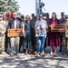 A group of military personnel and civilians gather for a memorial highway dedication ceremony honoring SFC Kyle Wehrly. Two participants hold brown highway signs reading 'SFC Kyle Wehrly Memorial Highway,' while a third person holds a photograph of the honoree in uniform. The event appears to take place at a military installation or memorial park, with a statue and flagpole visible in the background.