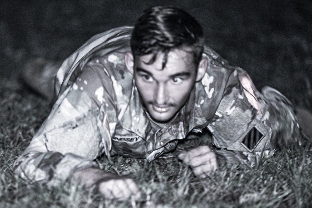 A young male in military camouflage uniform is photographed crawling on grass in a low-crawl position, looking directly at the camera. The black-and-white image appears to be taken at night or in low-light conditions. The subject appears to be engaged in a military training exercise.