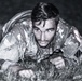 A young male in military camouflage uniform is photographed crawling on grass in a low-crawl position, looking directly at the camera. The black-and-white image appears to be taken at night or in low-light conditions. The subject appears to be engaged in a military training exercise.