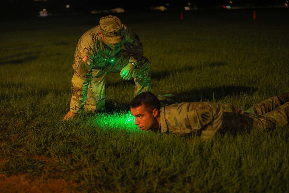 A nighttime military training exercise showing two soldiers in U.S. Army OCP (Operational Camouflage Pattern) uniforms on a grass field. One soldier is prone on the ground while a second soldier stands over him, illuminating the area with a green light source. The scene appears to depict a low-crawl drill or ground movement training exercise.