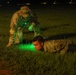 A nighttime military training exercise showing two soldiers in U.S. Army OCP (Operational Camouflage Pattern) uniforms on a grass field. One soldier is prone on the ground while a second soldier stands over him, illuminating the area with a green light source. The scene appears to depict a low-crawl drill or ground movement training exercise.