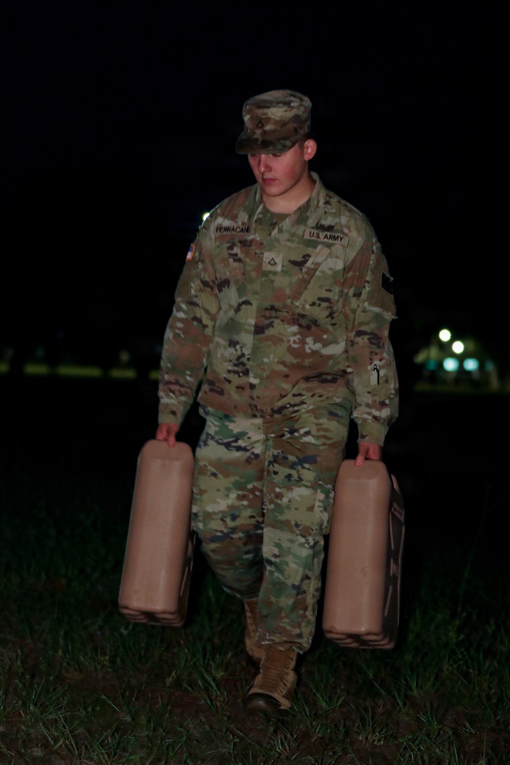 A U.S. Army soldier, identified by name tape as 'Ferracane,' is carrying two tan/beige military jerry cans or water containers at night. The soldier is wearing the Army Combat Uniform (ACU) in Operational Camouflage Pattern (OCP) with a patrol cap. The setting appears to be a grassy field or open area on a military installation, illuminated by artificial lighting.