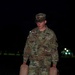 A U.S. Army soldier, identified by name tape as 'Ferracane,' is carrying two tan/beige military jerry cans or water containers at night. The soldier is wearing the Army Combat Uniform (ACU) in Operational Camouflage Pattern (OCP) with a patrol cap. The setting appears to be a grassy field or open area on a military installation, illuminated by artificial lighting.
