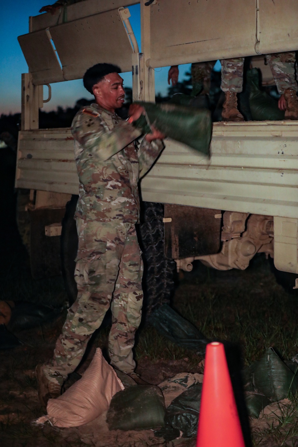 A soldier in US Army OCP (Operational Camouflage Pattern) uniform is loading or unloading green duffel bags or equipment bags onto/from a military tactical truck at dusk or dawn. Other soldiers are visible in the truck bed, appearing to assist with the transfer of gear. The scene appears to be a field operation or training exercise.