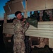 A soldier in US Army OCP (Operational Camouflage Pattern) uniform is loading or unloading green duffel bags or equipment bags onto/from a military tactical truck at dusk or dawn. Other soldiers are visible in the truck bed, appearing to assist with the transfer of gear. The scene appears to be a field operation or training exercise.