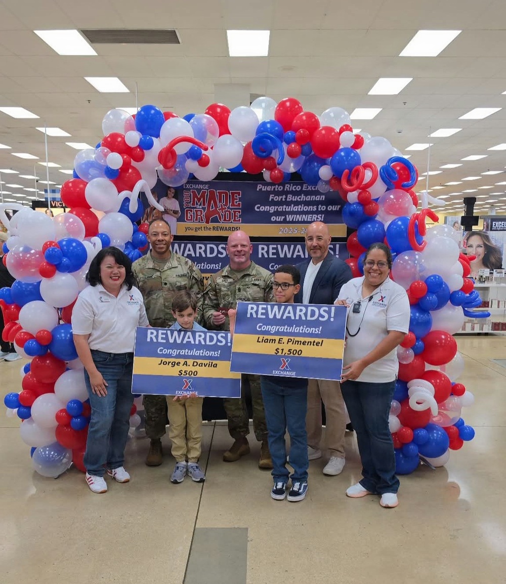 A rewards ceremony is taking place at the Puerto Rico Exchange at Fort Buchanan, where two winners are being recognized. Jorge A. Davila is receiving a $500 reward and Liam E. Pimentel is receiving a $1,500 reward from the Exchange 'You Made the Grade' program. The group poses in front of a red, white, and blue balloon arch inside a military exchange store.