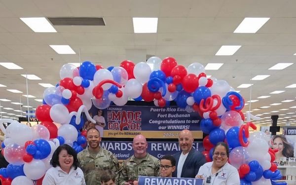 A rewards ceremony is taking place at the Puerto Rico Exchange at Fort Buchanan, where two winners are being recognized. Jorge A. Davila is receiving a $500 reward and Liam E. Pimentel is receiving a $1,500 reward from the Exchange 'You Made the Grade' program. The group poses in front of a red, white, and blue balloon arch inside a military exchange store.
