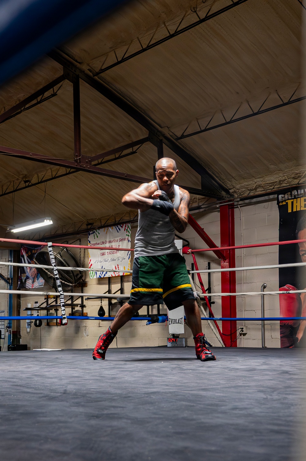 A male boxer is training inside a boxing ring at an indoor gym, shown in a defensive stance with hand wraps on his fists. He is wearing a gray tank top, green and yellow shorts, and red and black boxing boots. The gym features Everlast-branded ropes, a speed bag, banners on the walls, and exposed steel roof trusses.