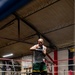 A male boxer is training inside a boxing ring at an indoor gym, shown in a defensive stance with hand wraps on his fists. He is wearing a gray tank top, green and yellow shorts, and red and black boxing boots. The gym features Everlast-branded ropes, a speed bag, banners on the walls, and exposed steel roof trusses.
