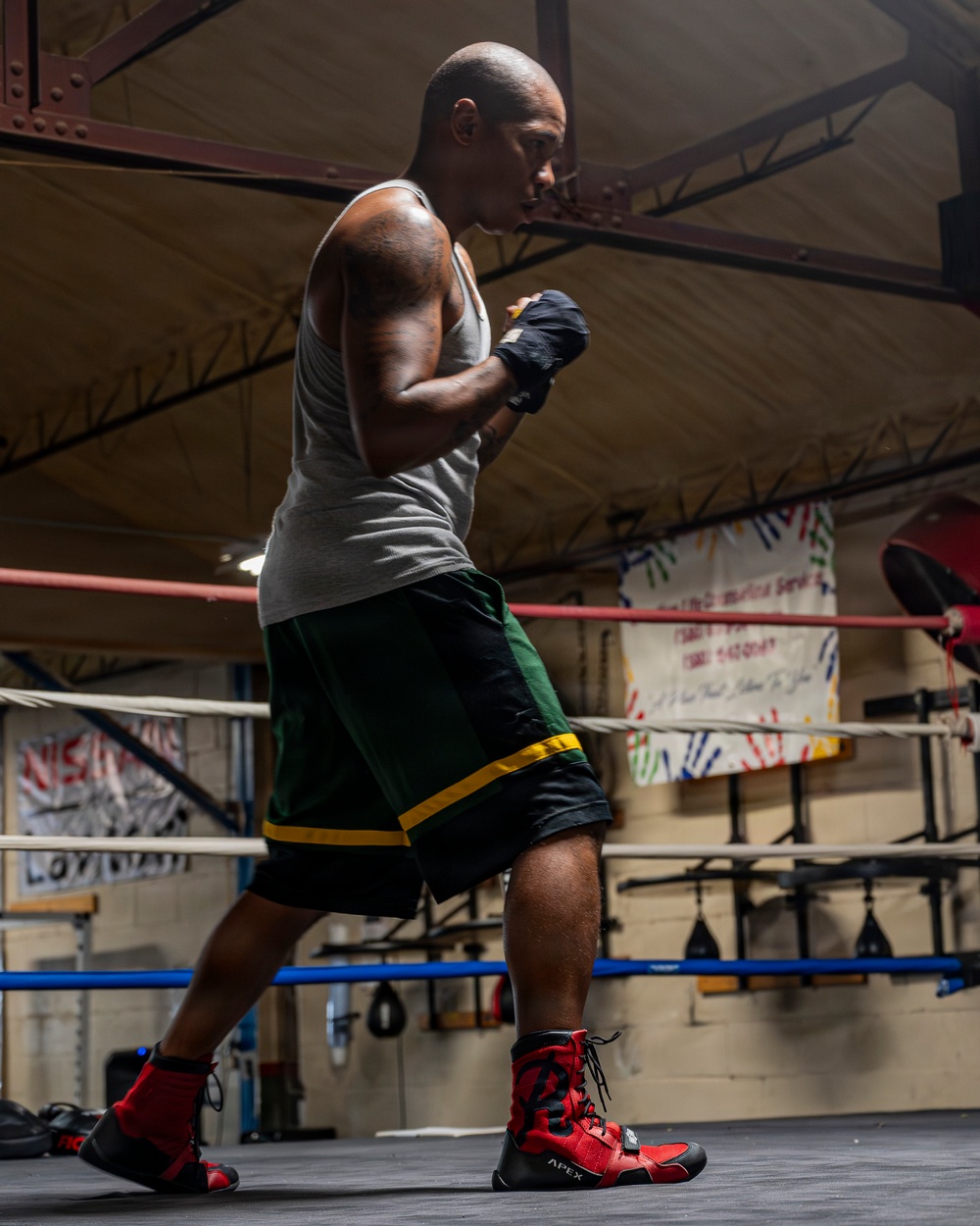 A muscular, bald male boxer is training inside a boxing gym, standing in a fighting stance within a boxing ring. He is wearing hand wraps, a gray tank top, green and black shorts with yellow trim, and red-and-black high-top boxing boots branded 'APEX'. The gym environment includes ring ropes, hanging banners, and training equipment visible in the background.