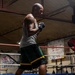A muscular, bald male boxer is training inside a boxing gym, standing in a fighting stance within a boxing ring. He is wearing hand wraps, a gray tank top, green and black shorts with yellow trim, and red-and-black high-top boxing boots branded 'APEX'. The gym environment includes ring ropes, hanging banners, and training equipment visible in the background.