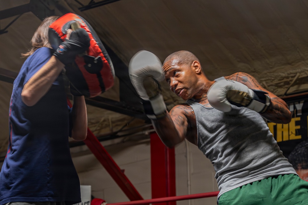 A heavily tattooed male boxer wearing gray tank top and green shorts is training in a boxing gym, throwing punches at focus mitts held by a trainer. The session is taking place inside a boxing ring with red corner posts visible in the background. The image captures the intensity and motion of a pad work training session.