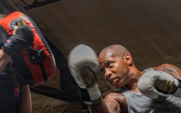 A heavily tattooed male boxer wearing gray tank top and green shorts is training in a boxing gym, throwing punches at focus mitts held by a trainer. The session is taking place inside a boxing ring with red corner posts visible in the background. The image captures the intensity and motion of a pad work training session.