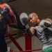 A heavily tattooed male boxer wearing gray tank top and green shorts is training in a boxing gym, throwing punches at focus mitts held by a trainer. The session is taking place inside a boxing ring with red corner posts visible in the background. The image captures the intensity and motion of a pad work training session.