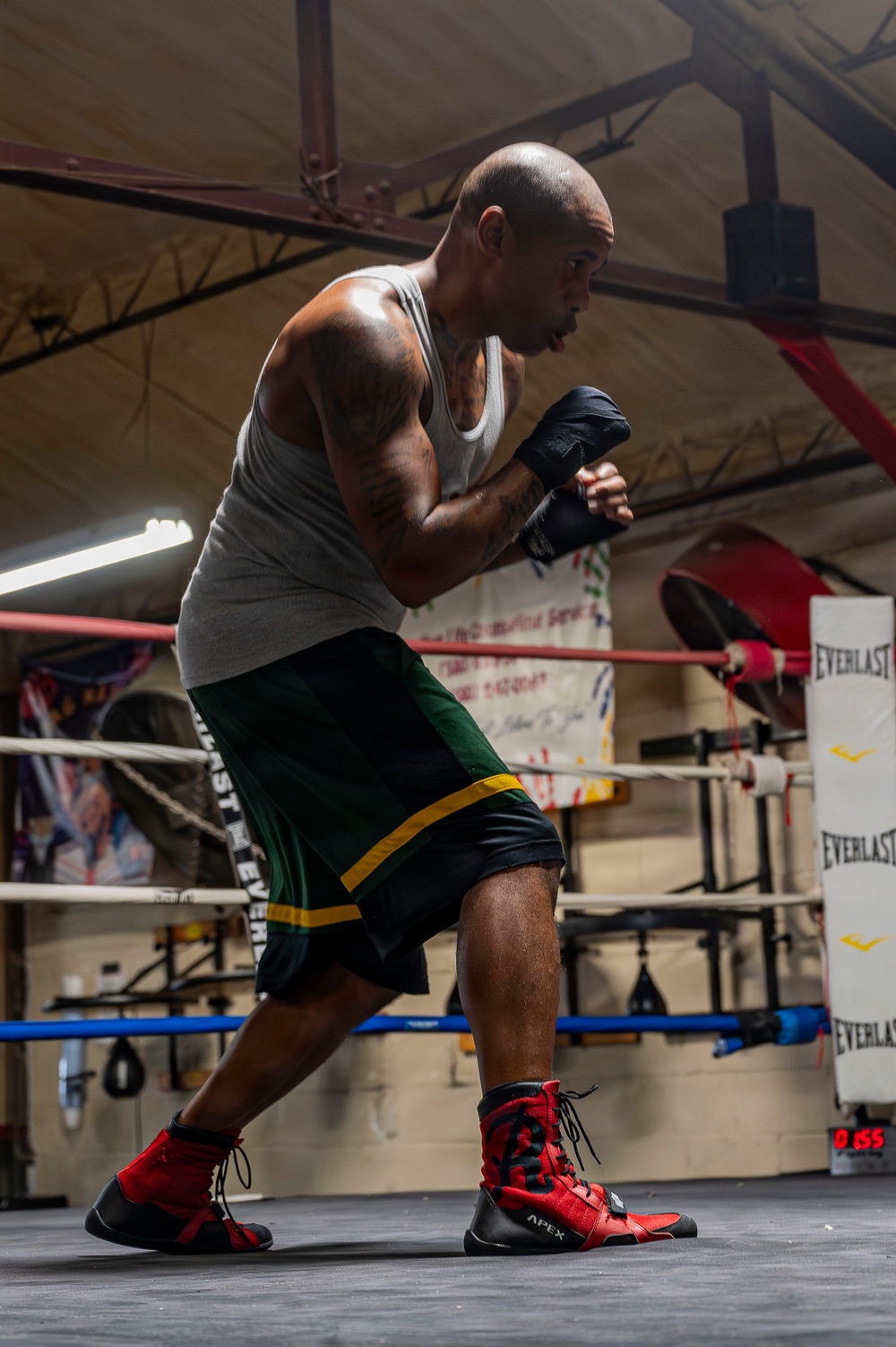 A bald male boxer is captured mid-training stance inside a boxing gym, wearing hand wraps and boxing shoes in a fighting guard position. He is standing inside or near a boxing ring equipped with Everlast ropes and equipment. The image is well-lit with dramatic side lighting that highlights his muscular physique and athletic posture.