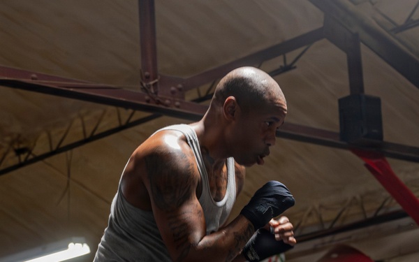 A bald male boxer is captured mid-training stance inside a boxing gym, wearing hand wraps and boxing shoes in a fighting guard position. He is standing inside or near a boxing ring equipped with Everlast ropes and equipment. The image is well-lit with dramatic side lighting that highlights his muscular physique and athletic posture.