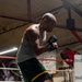 A bald male boxer is captured mid-training stance inside a boxing gym, wearing hand wraps and boxing shoes in a fighting guard position. He is standing inside or near a boxing ring equipped with Everlast ropes and equipment. The image is well-lit with dramatic side lighting that highlights his muscular physique and athletic posture.